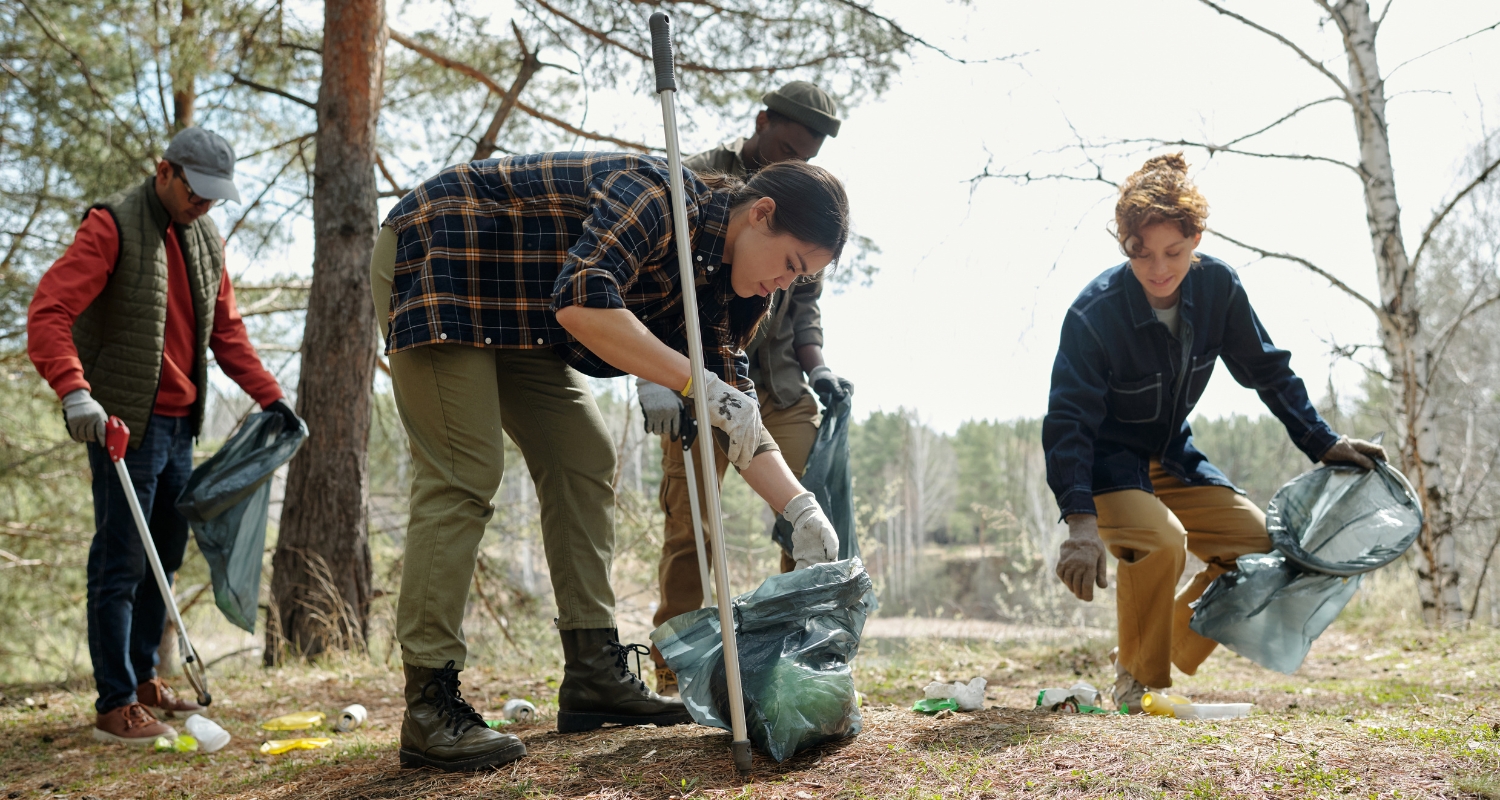 Gruppo di giovani volontari raccoglie rifiuti in un’area verde boschiva utilizzando pinze e sacchi, durante un’attività di cura dell’ambiente all’aperto.
