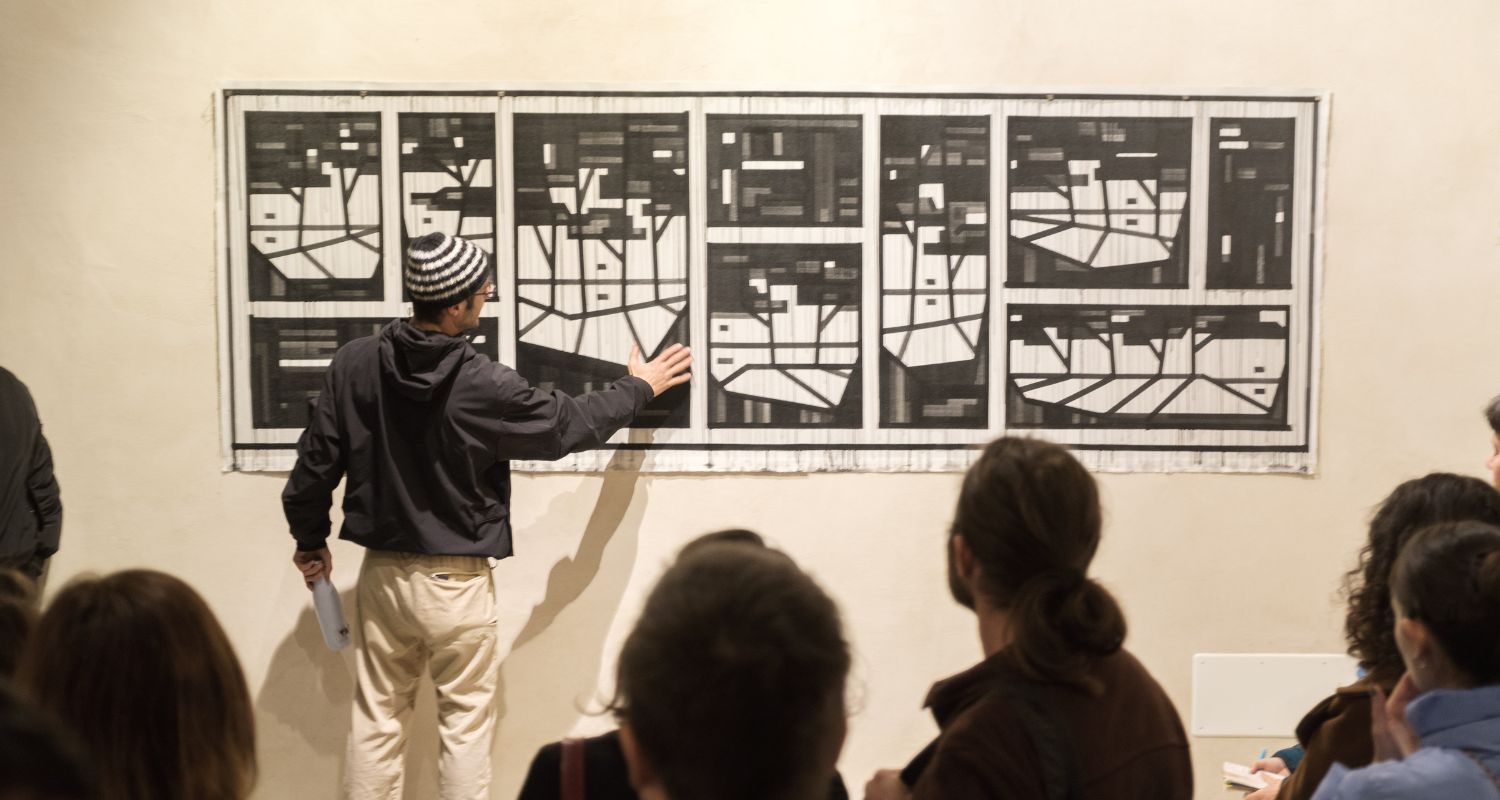 Una persona presenta un'opera artistica astratta in bianco e nero composta da pannelli geometrici appesi a una parete, mentre un gruppo di visitatori osserva la spiegazione durante una visita in una galleria d'arte.