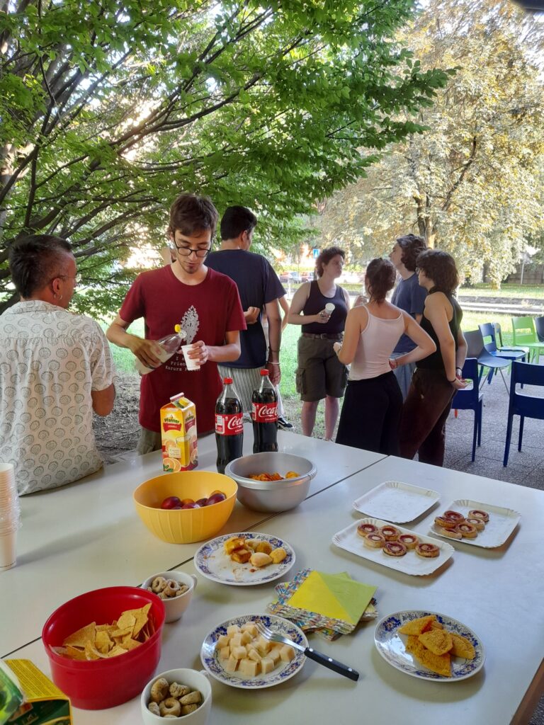 Un gruppo di giovani fa conversazione durante un aperitivo. In primo piano c'è un tavolo con pizzette, salatini e bibite.