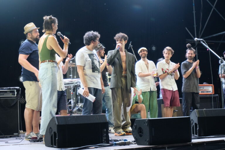 Un gruppo di ragazzi è su un palco. Uno di loro sta parlando al microfono guardano una ragazza, anche lei sul palco con un microfono in mano.