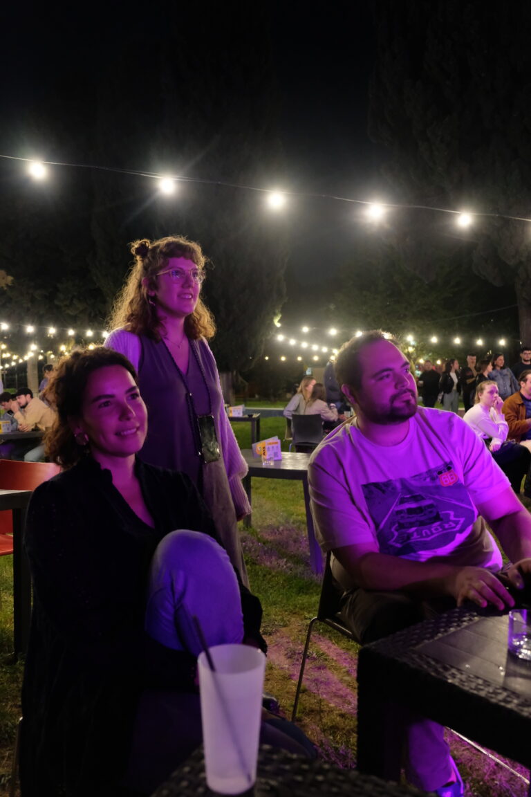 Tre persone sono sedute a un tavolo e guardano davanti a sé, nella stessa direzione, illuminate da un fascio di luce viola che probabilmente proviene da un palco.