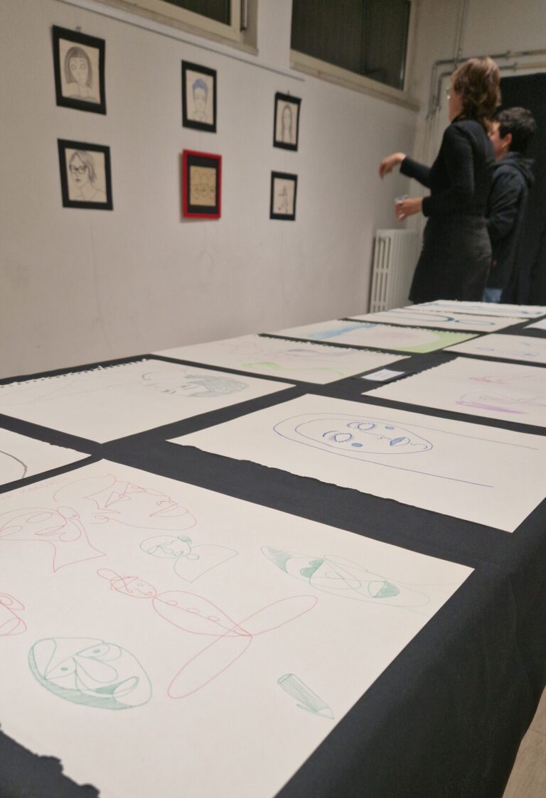 Scorcio di una mostra artistica: in primissimo piano una serie di disegni, per lo più ritratti molto stilizzati, è disposta su un tavolo con una tovaglia nera. Sullo sfondo due ragazze stanno osservando e commentando dei quadri appesi alla parete.