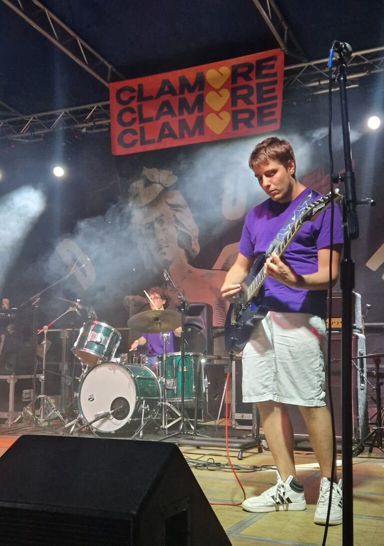 Due ragazzi suonano su un palco. Sullo sfondo il batterista, in primo piano il bassista. Entrambi indossano la stessa maglietta di colore viola.