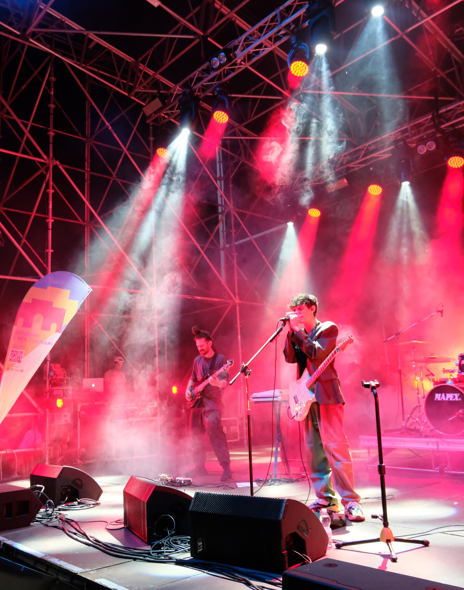 Due musicisti suonano su un palco, illuminato da luci rosse: uno di loro suona il basso, l'altro canta reggendo il microfono con due mani.