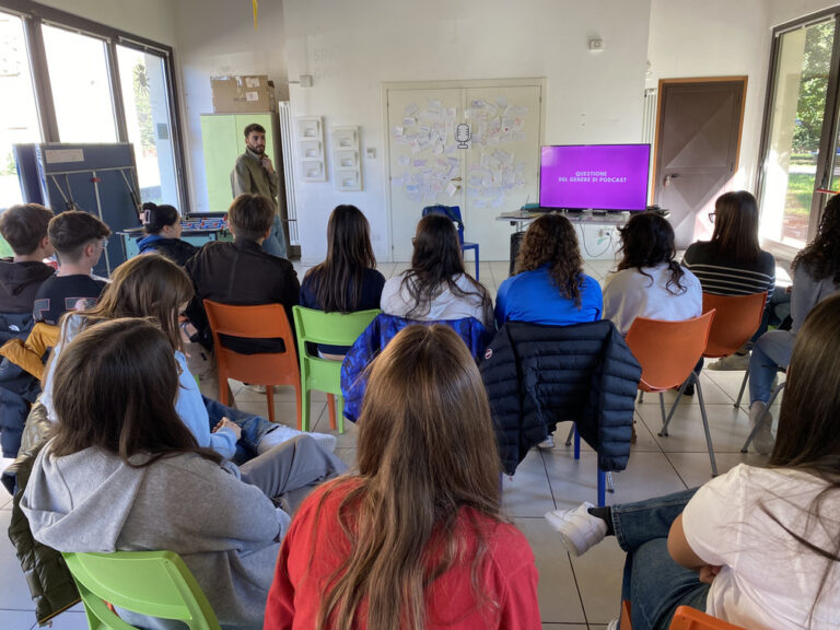 Un gruppo di studenti è fotografato di spalle, seduto a semicerchio. Un ragazzo è in piedi davanti a loro e parla aiutandosi con delle slide proiettate su una tv.