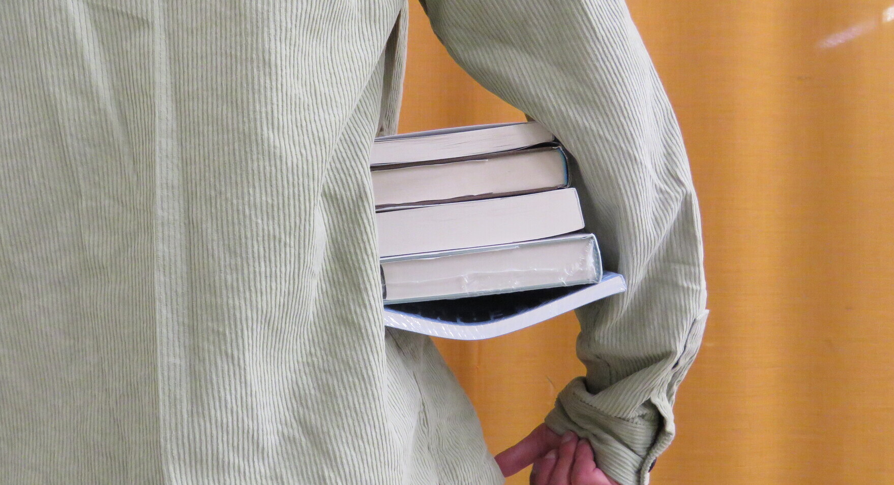Un ragazzo con una camicia di velluto verde chiaro regge una pila di libri sotto il braccio. L'inquadratura è stretta sul dettaglio dei libri e del braccio che li tiene.