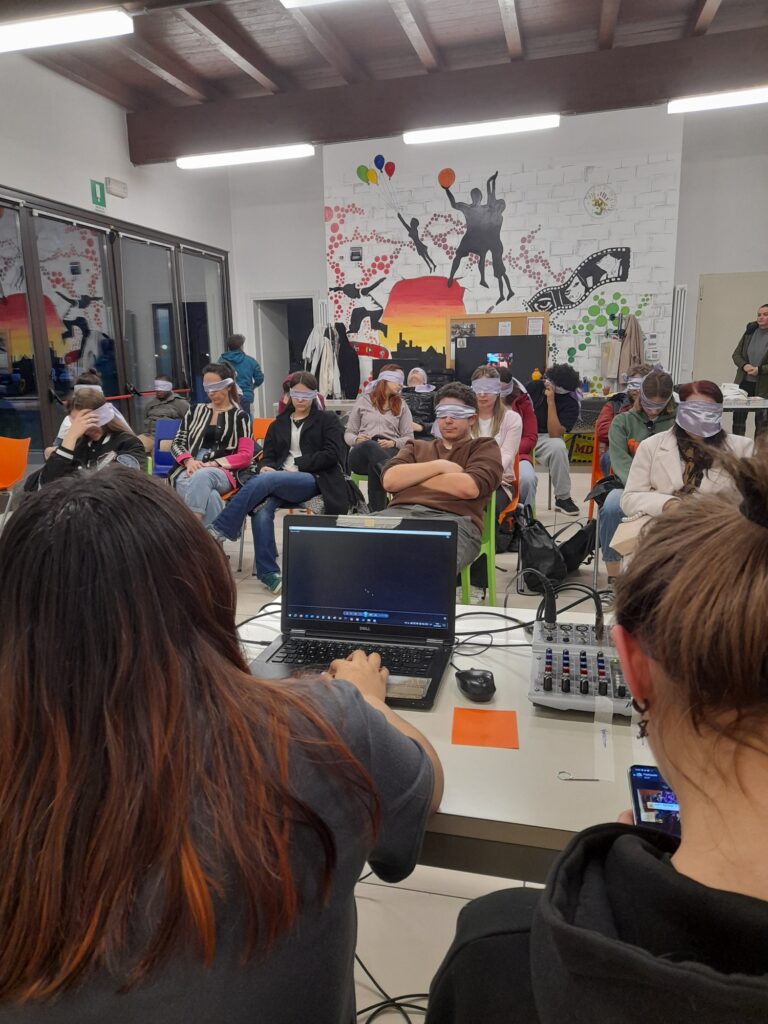 Un gruppo di persone è seduto in file e indossano delle bende sugli occhi. Due ragazze in primo paino, fotografate di spalle, stanno facendo ascoltare una traccia audio dal pc.