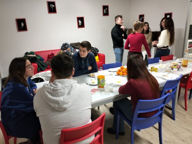 Un gruppo di giovani parla e scherza seduto a una lunga tavolata apparecchiata con piatti e bicchieri di carta. Un altro piccolo gruppo sta facendo conversazione in piedi, alle spalle del tavolo.