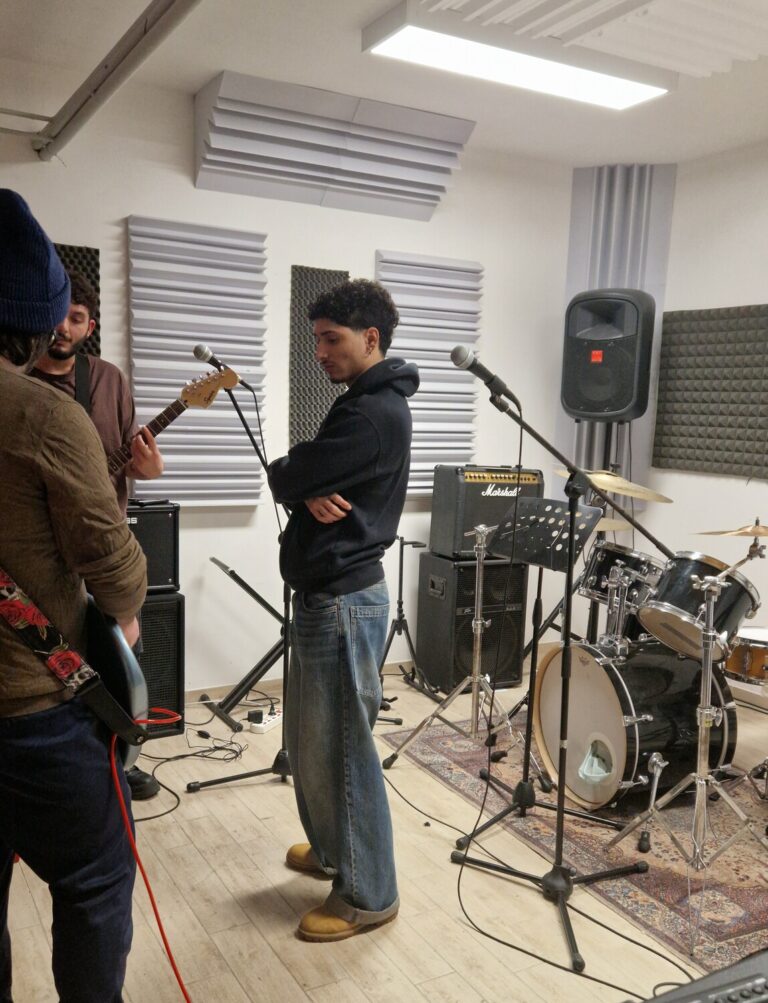 Tre ragazzi si stanno esercitando in una sala prove musicale. Due di loro stanno accordando il basso e la chitarra, il terzo li guarda.