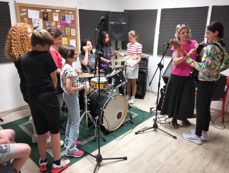 Un gruppo di sette ragazze e un ragazzo sta provando le strumentazioni di una sala prove musicale. Una donna sta aiutando una ragazza a regolare la cinghia di una chitarra elettrica, mentre il resto del gruppo le guarda.