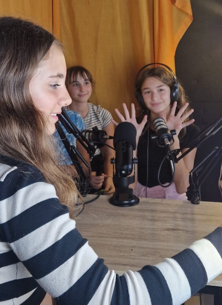 Tre ragazze sedute a un tavolo, provano dei microfoni da studio. Una quarta ragazza, in primo piano, sembra stia per far partire una registrazione.