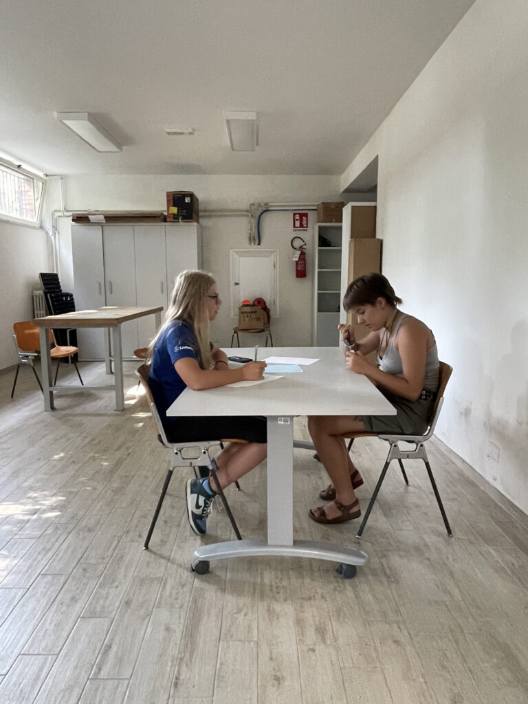 Due ragazze sono sedute a un tavolo. Una sta tagliando un pezzo di scotch con le forbici, l'altra la guarda tenendo in mano una penna, come fosse sul punto di scrivere qualcosa s un foglio.