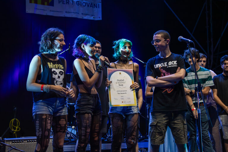 Un gruppo di tre ragazze e un ragazzo è su un palco, illuminato da una luce blu. Una di loro regge l'attestato di un premio, un'altra sta parlando al microfono. Indossano pantaloncini di jeans e magliette di gruppi punk. Sullo sfondo ci sono altri ragazzi, più in disparte.