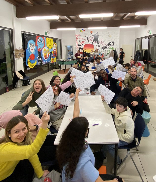 Un gruppo di persone molto numeroso siede a una lunga tavolata. Guardano in camera, sorridono e sventolano le cartelle della tombola.