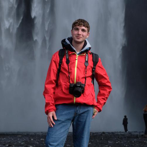 Andrea è in posa di fronte a delle cascate. Sorride, ha la mano su un fianco, indossa una giacca rossa e ha al collo la macchina fotografica.