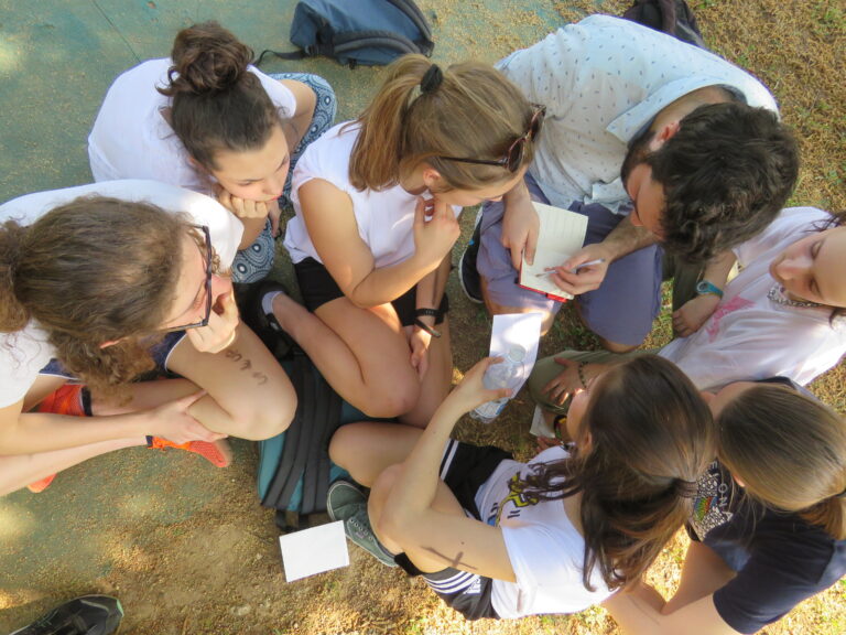 Un gruppo di ragazze è seduto per terra, riunito attorno a un ragazzo che scrive qualcosa su un quadernino. La foto è scattata dall'alto.