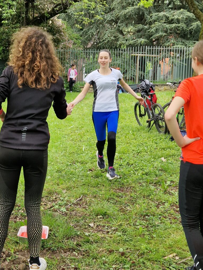 Una ragazza sta correndo verso un'altra tendendole la mano, come a passarle una staffetta. La seconda ragazza è protesa verso la prima, pronta a scattare. L'ambientazione è quella di un parco, con delle biciclette appoggiate al cavalletto poco più avanti.