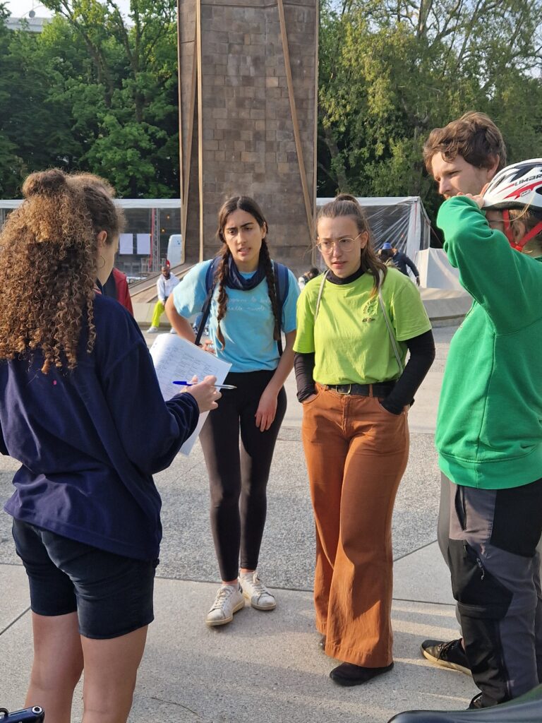 Un gruppo di giovani guarda una ragazza riccia, fotografata di spalle, che sta leggendo loro qualcosa scritto su un foglio che regge in mano.