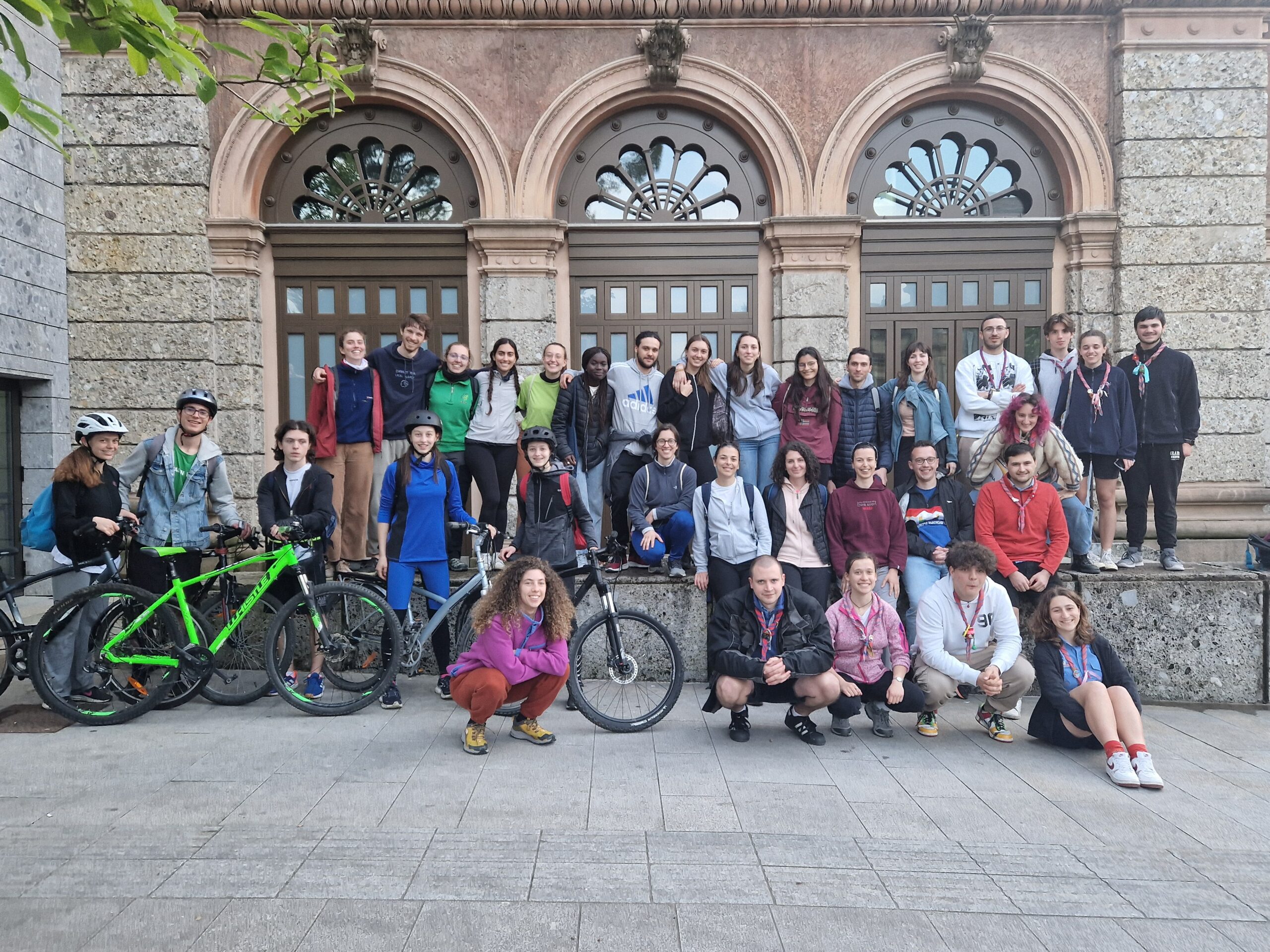 Foto di gruppo di un gruppo numeroso di giovani in abiti sportivi. Alcune ragazze in prima fila reggono delle biciclette e indossano dei caschetti La foto è scattata all'aperto, di fronte a un edificio storico.