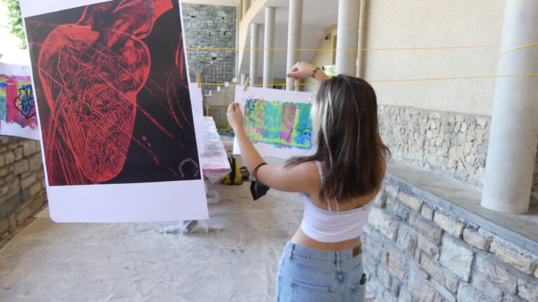 Una ragazza è fotografata di spalle mentre appende con delle mollette dei poster e delle stampe a un filo, teso tra due colonne in un cortile.
