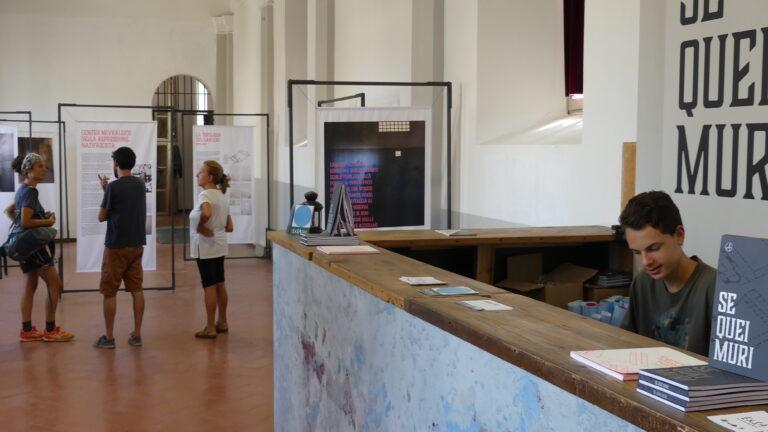 Sullo sfondo un gruppo di persone sta visitando una mostra, leggendo dei totem esplicativi. In primo piano, un ragazzo è seduto dietro al bancone dell'accoglienza.
