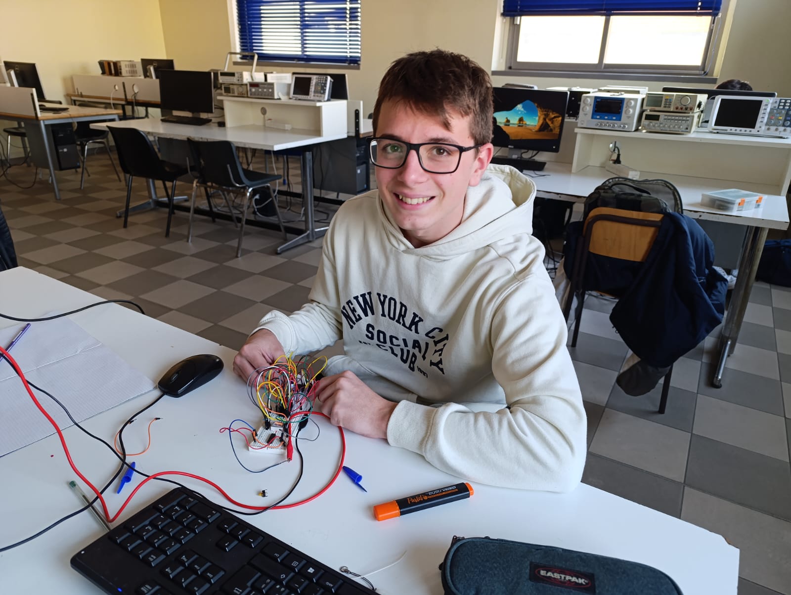 Andrea sorride in aula con una felpa chiara. Ha gli occhiali e sta lavorando su una serie di cavi elettrici