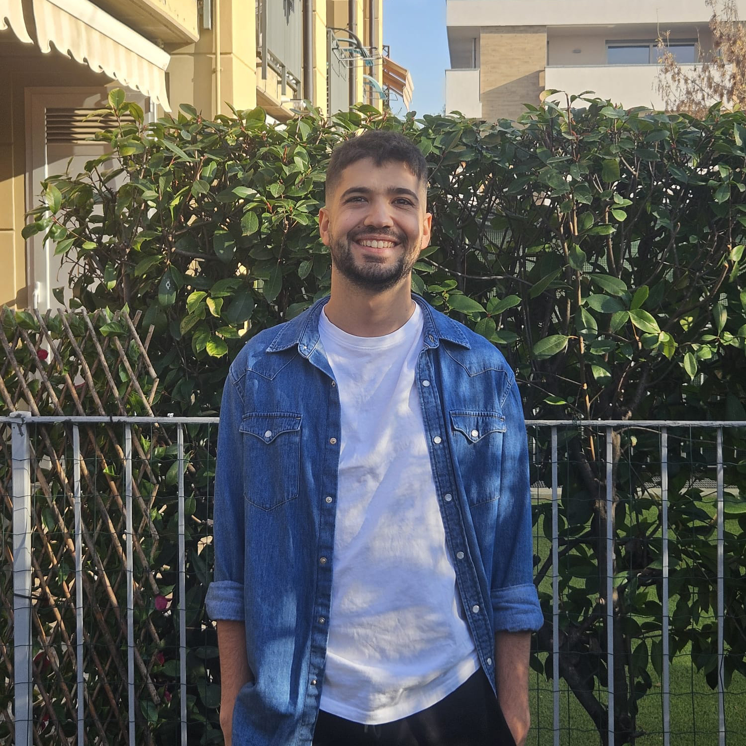 Fabio in uno spazio verde, davanti a un cancelletto. Sorride, indossa una camicia di jeans e una t-shirt bianca