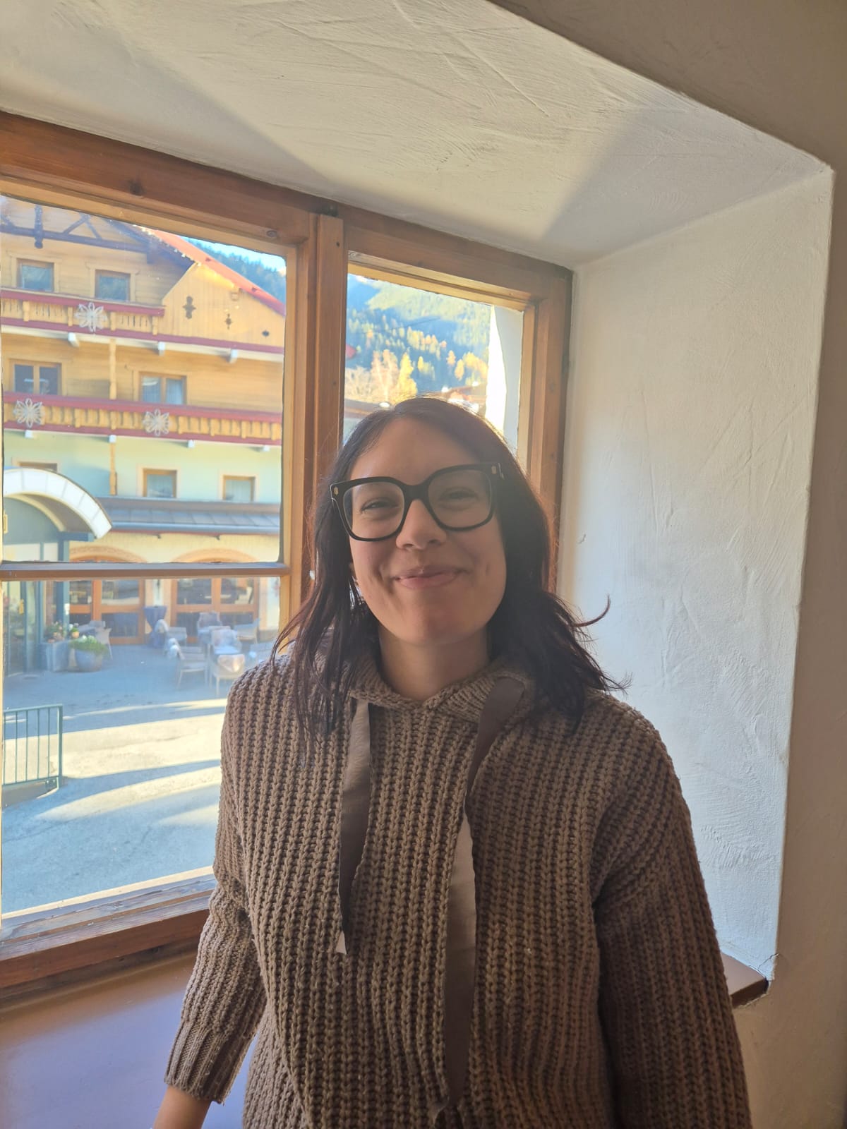 Nicole accanto a una finestra. Sorride e ha un maglione beige. Porta gli occhiali