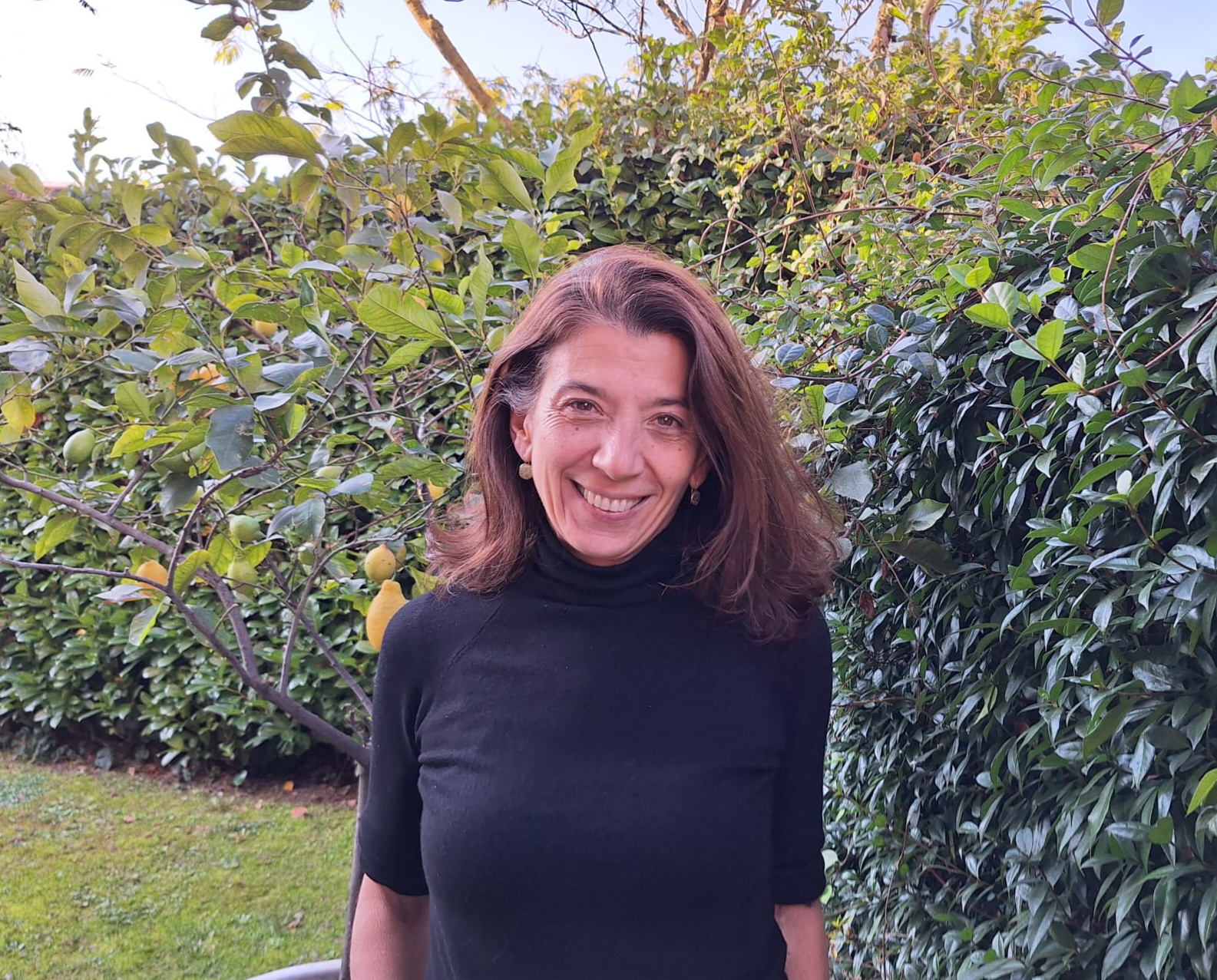 Giovanna in questa foto è in mezzo al verde, accanto a una siepe. Sorride, ha una maglia scura.