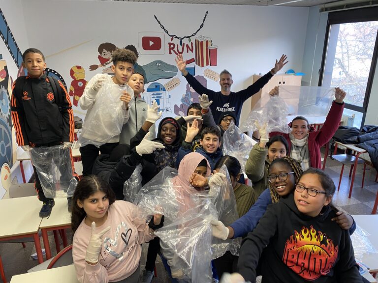 Un gruppo di ragazze e ragazzi sui di circa 11-12 anni posa in una aula scolastica, mostrando delle buste di plastica vuote. Insieme a loro ci sono due persone adulte, una delle quali indossa una felpa blu con il logo dell'associazione PlasticFree.