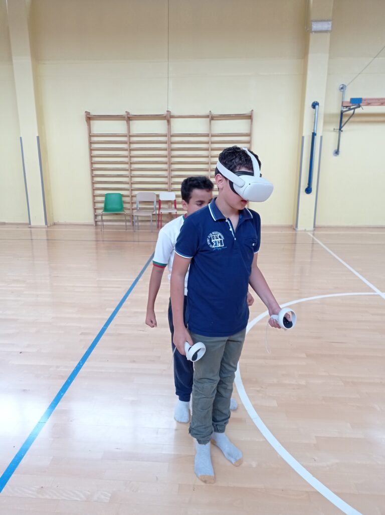 Un ragazzo indossa un visore VR e tiene in mano due manopole controller, mentre un altro dietro di lui sembra copiarne i movimenti. La foto è stata scattata dentro una palestra con pavimento in parquet.