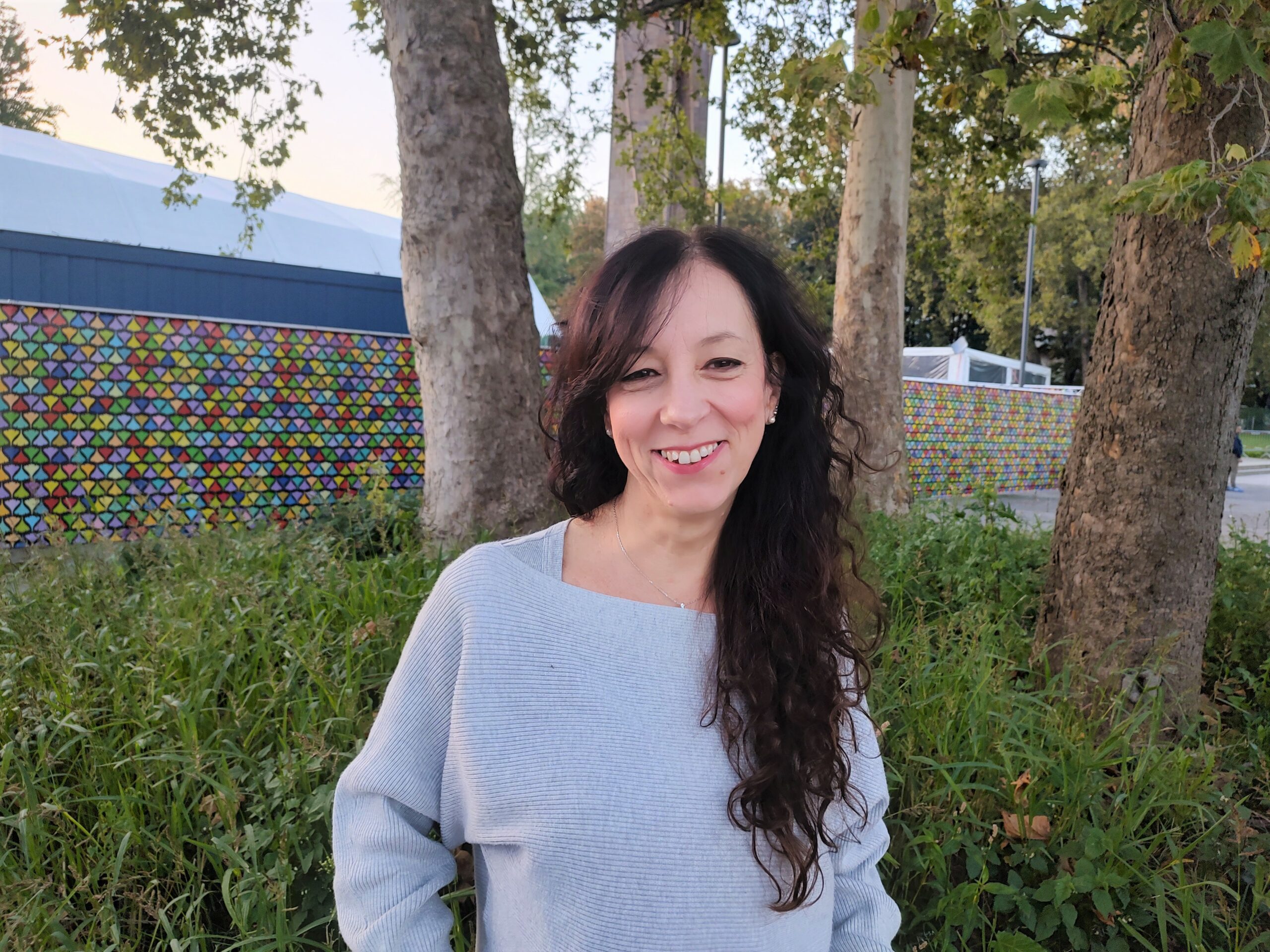 In questa foto Nadia è all'aperto, dietro di lei alcuni alberi. Sorride, ha i capelli lunghi e scuri, porta un maglione chiaro