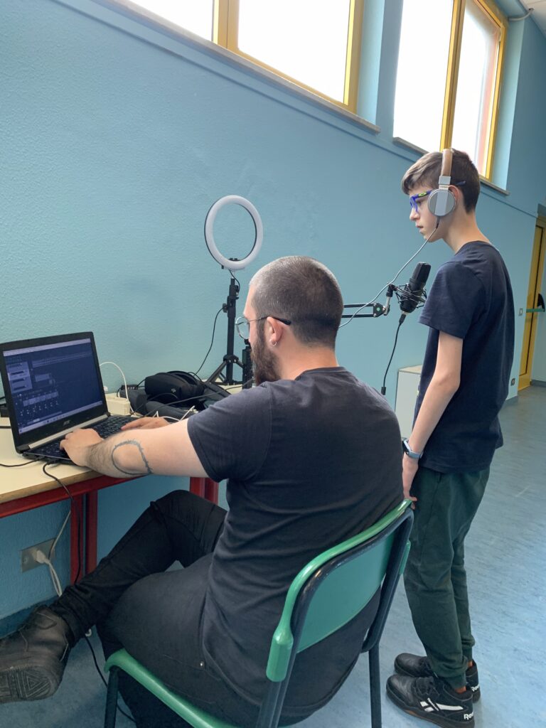 Un ragazzo è in piedi davanti a un microfono e indossa delle cuffie. Un educatore, seduto accanto a lui, gli sta mostrando sul pc un programma per registrare tracce audio.