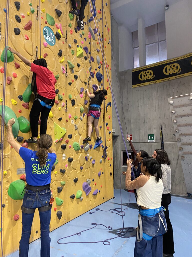 Un gruppo di ragazze sta provando a scalare una parete da arrampicata in palestra. Sono tenute in sicurezza con le corde da altre ragazze a terra e guidate da un istruttore.