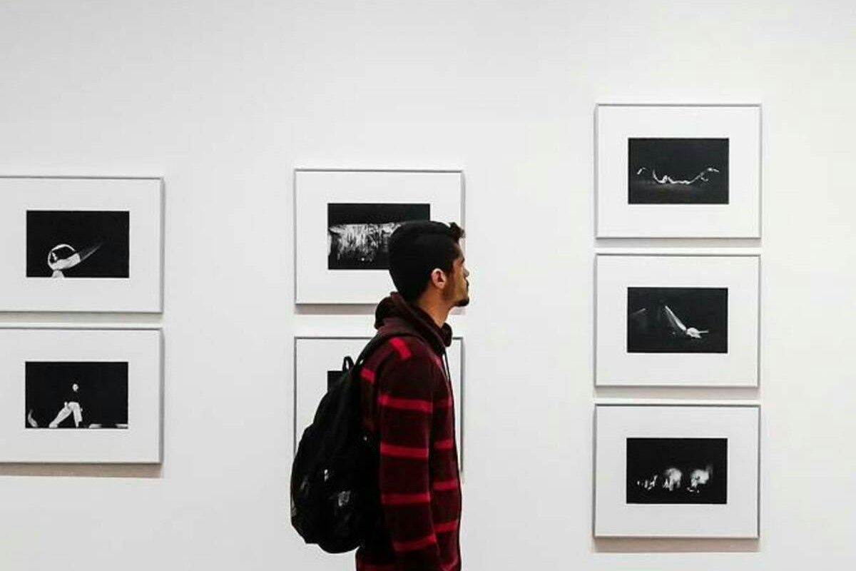 Un ragazzo con una felpa a righe e uno zaino sulle spalle è a una mostra d'arte contemporanea e osserva quelle che sembrano delle fotografie, incorniciate sulla parete.