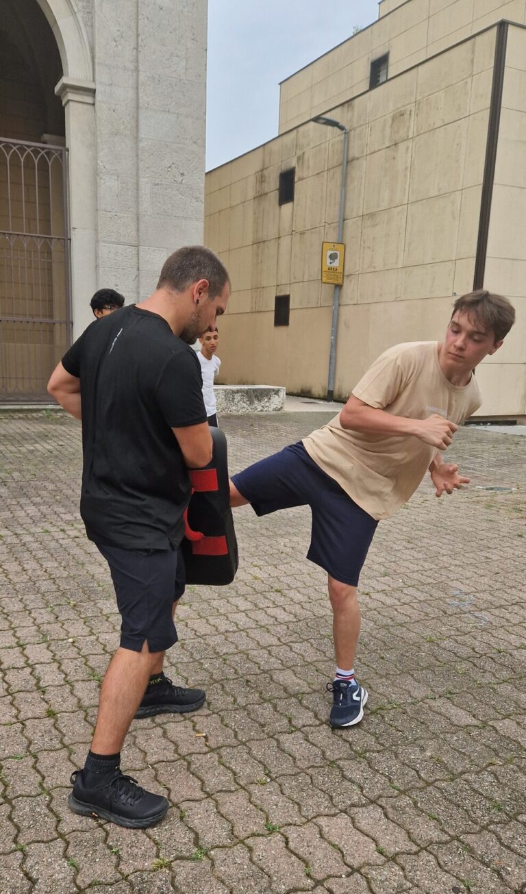 Un ragazzo si sta esercitando a tirare calci, che l'istruttore di boxe para con un paracolpi. La foto è scattata all'aperto, in un piazzale.