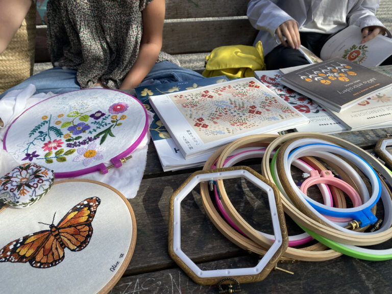 Sopra il tavolo in legno di un parco sono appoggiati strumenti per il ricamo: dei piccoli telai, libri con schemi, aghi e un paio di lavori già iniziati. Due giovani sullo sfondo sfogliano dei libri con gli schemi.