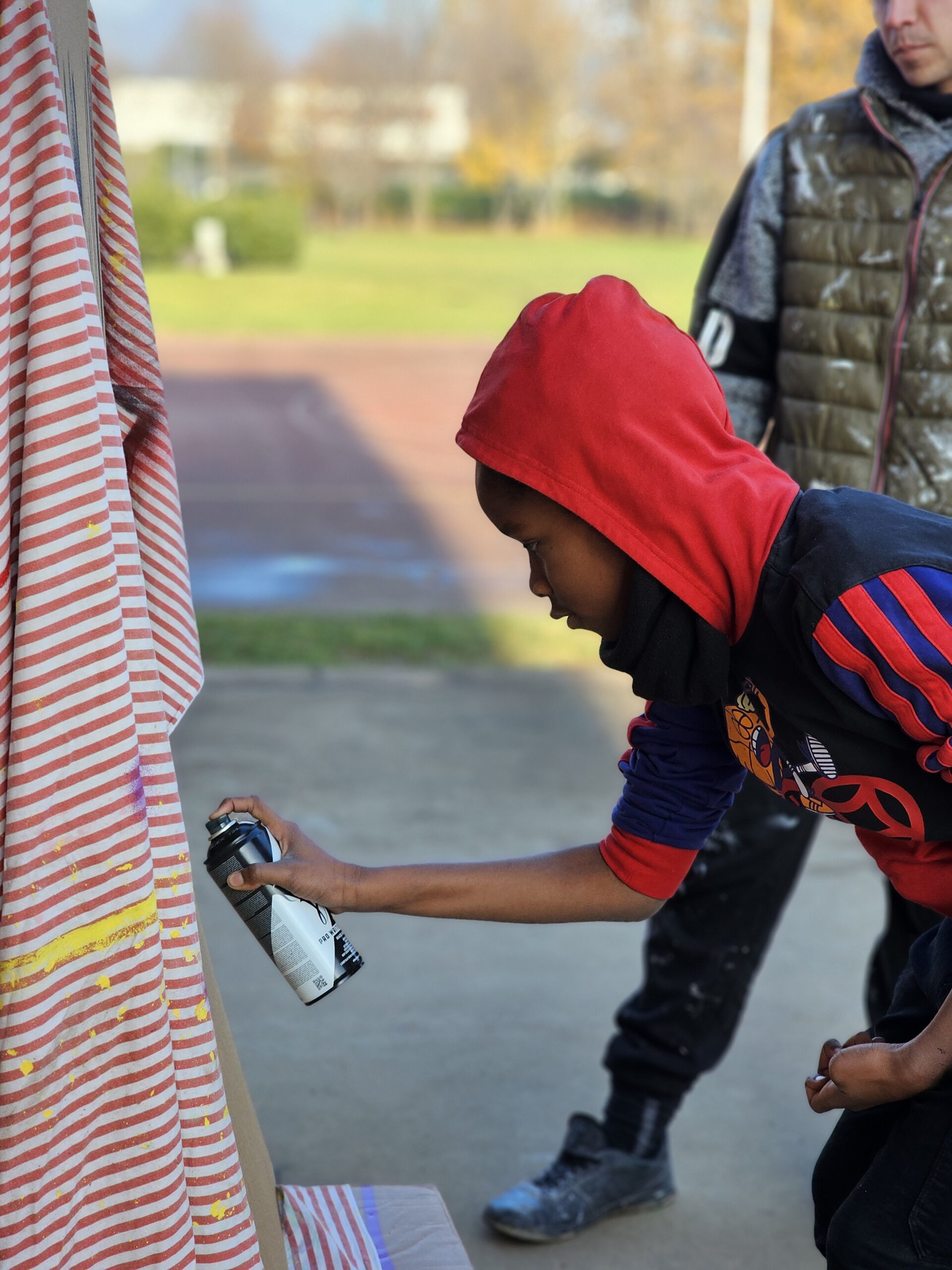 Un ragazzino si sta esercitando a usare una bomboletta spray su un telo di stoffa a righe. Indossa un felpa con il cappuccio rosso tirato su.