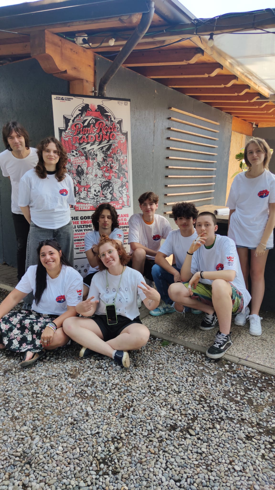 Un gruppo giovani posa sorridente davanti al totem della settima edizione del Punk Rock Raduno. La maggior parte di loro indossa una maglietta bianca con scritto 