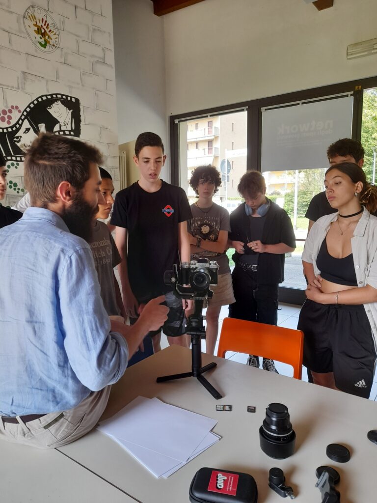 Un ragazzo - il regista - sta mostrando come utilizzare una videocamera a un gruppo di ragazzi e ragazze più giovani, che osservano con attenzione.