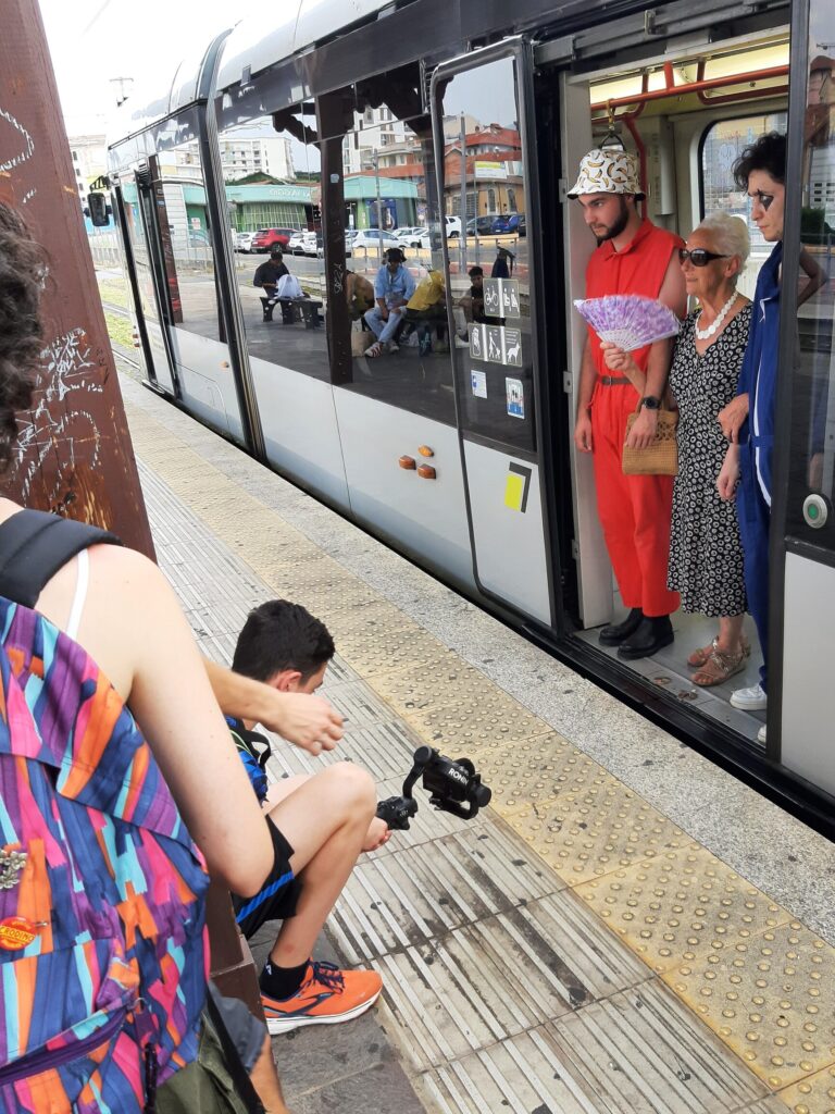 Una signora anziana sta scendendo dal tram, sorretta ai lati da due ragazzi in tuta da lavoro. Un ragazzo è sulla banchina e filma la scena dal basso.