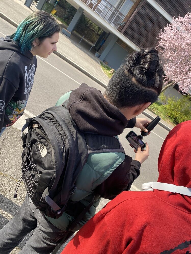 Un ragazzo fotografato di spalle sta facendo delle riprese con una mini-cam. Una ragazzo e un ragazzo ai suoi due lati lo osservano riprendere.