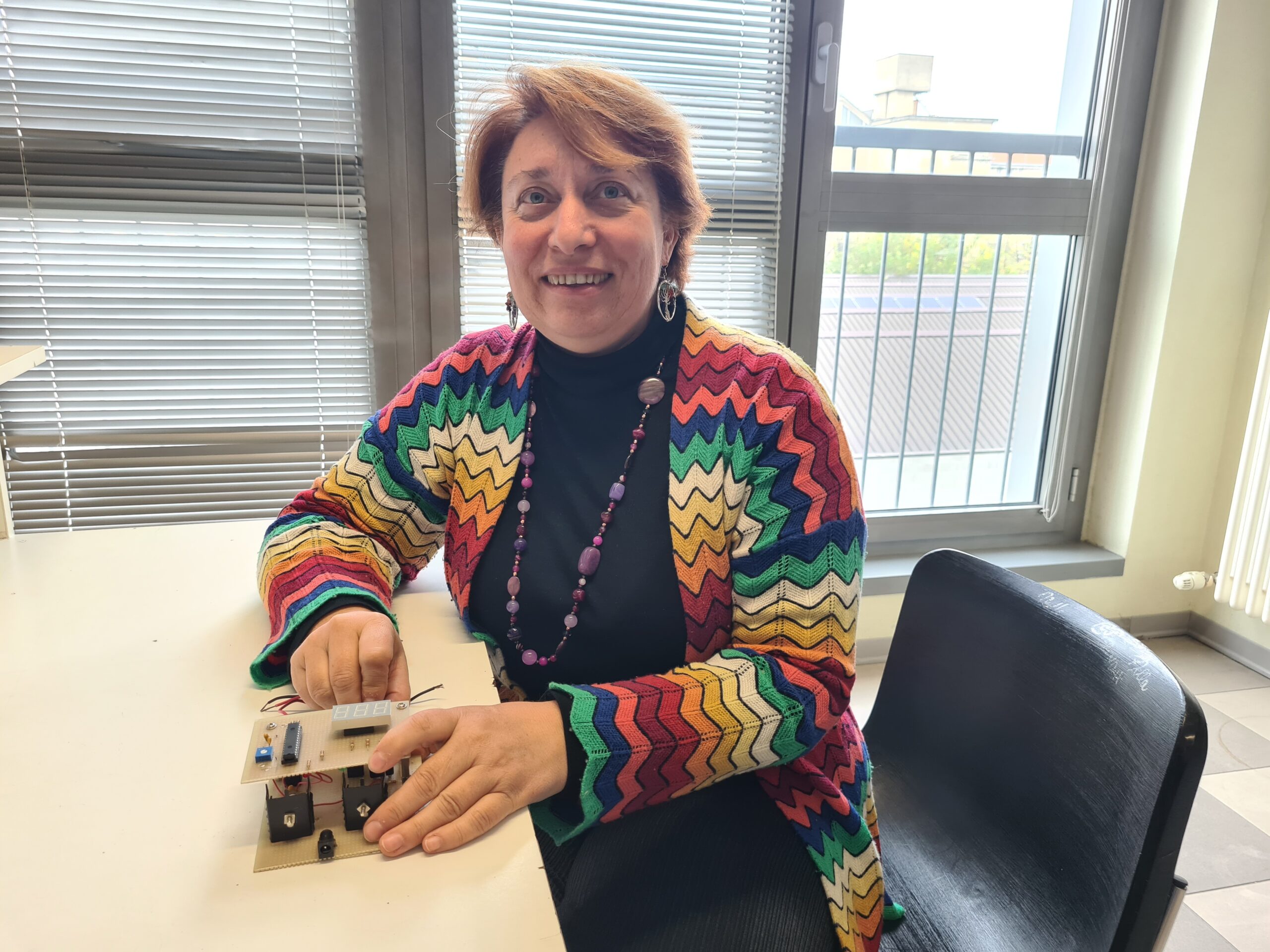 Vera ha un maglione colorato, sorride guardando in camera mentre tra le mani tiene una scheda elettronica