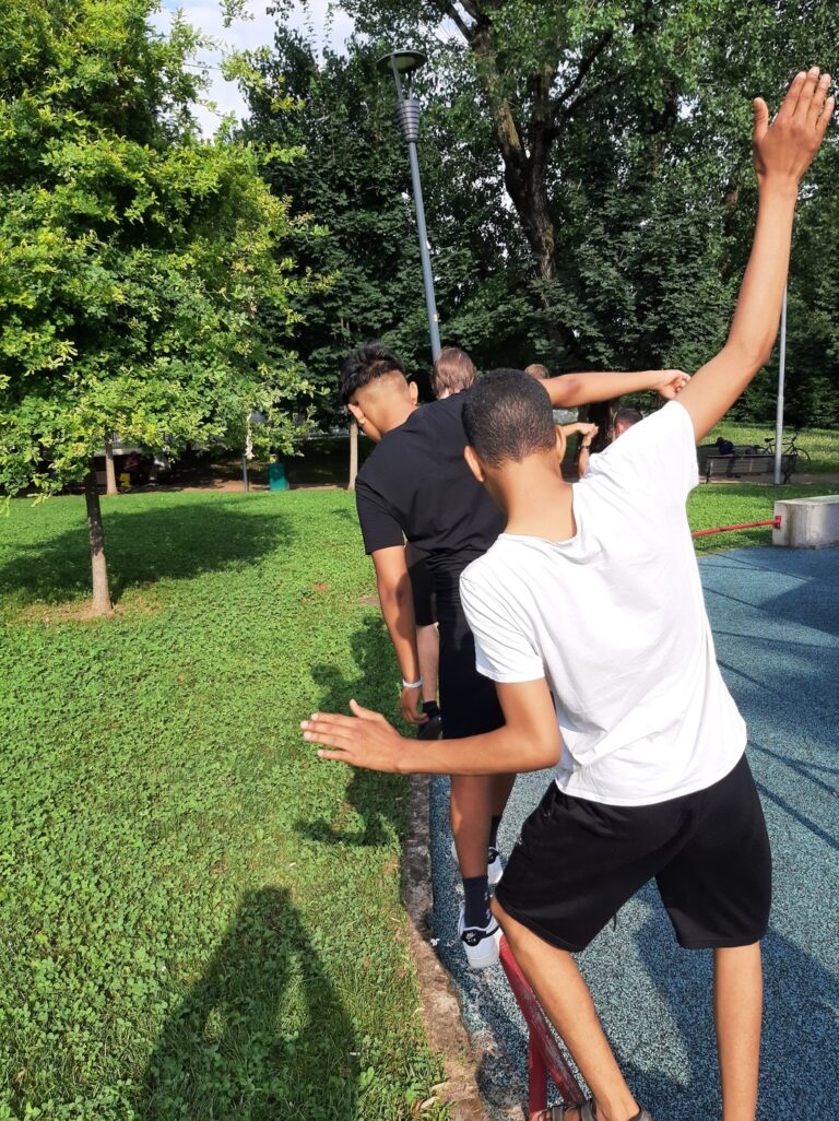 Un gruppo di ragazzi sta camminando in fila indiana su una sbarra di metallo rossa, a pochi centimetri da terra, cercando di mantenere l'equilibrio. La foto è stata scattata in un parco in una giornata di sole.
