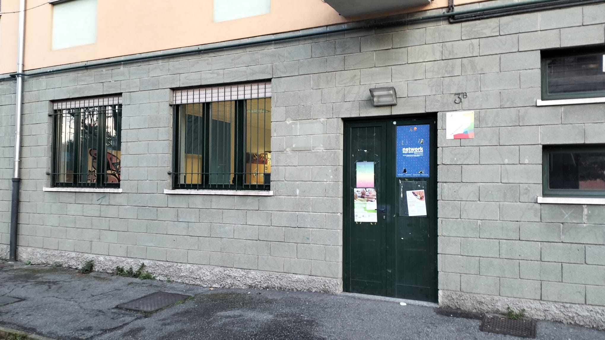 La facciata dello Spazio per Giovani nel quartiere di Grumello. È un edificio con i mattoni grigi e una porta verde dove sono attaccati poster delle iniziative dello Spazio.