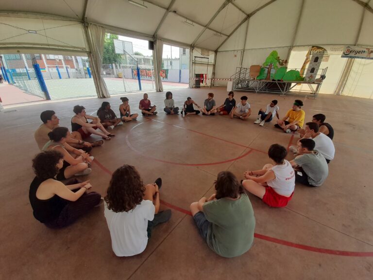 Un gruppo di giovani è fotografato in cerchio all'interno di una palestra. Siedono a gambe incrociate.