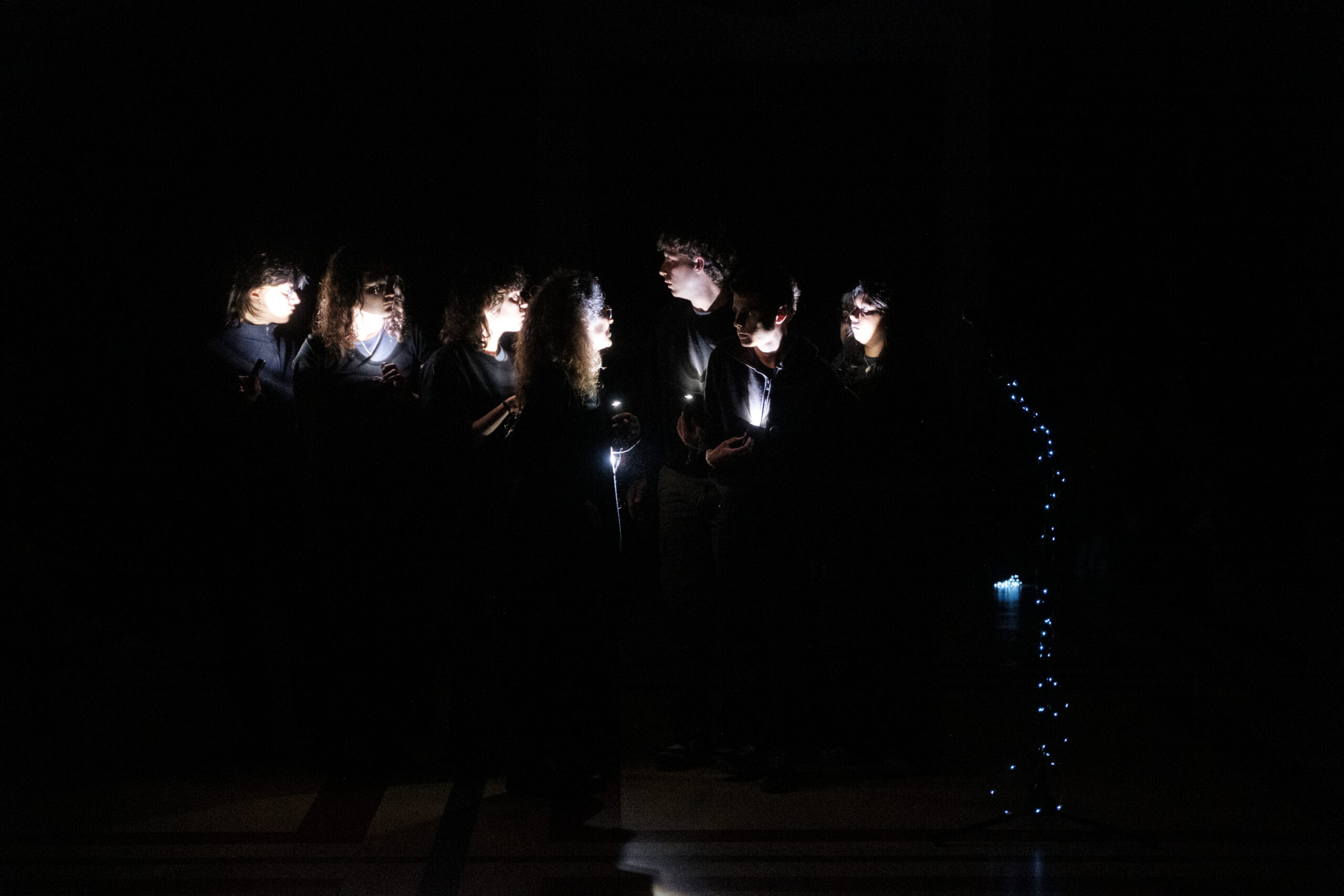 Un gruppo di giovani è fotografato durante una performance. L'ambiente è completamente al buio, tranne che per delle torce che tengono in mano e si puntano contro il viso.