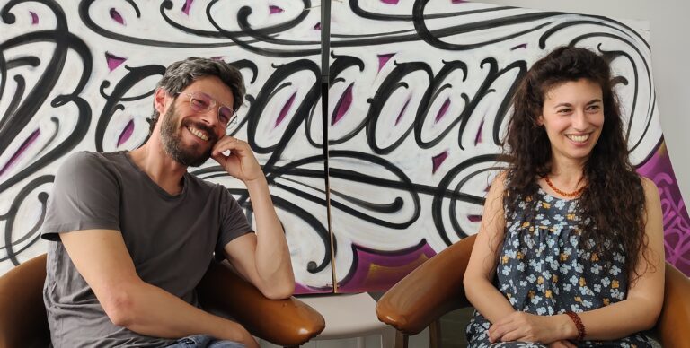 Fotografia di una Job Talk. Giorgio, consulente Informagiovani, è seduto a sinistra, l'ospite è a destra. Sorridono, hanno abiti leggeri e sullo sfondo c'è un graffito