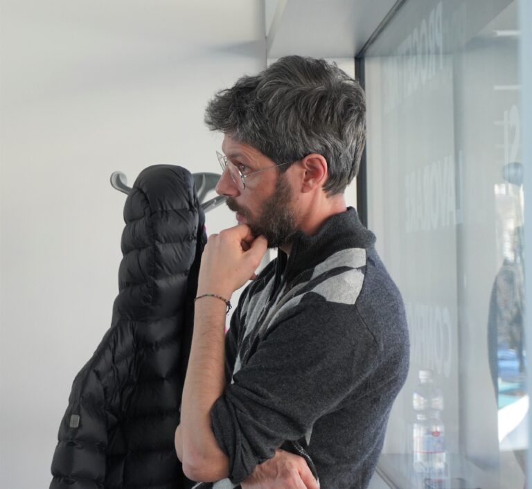 Consulente di orientamento dell'Informagiovani durante la giornata del placement. È appoggiato con la schiena a una finestra, mentre ascolta con il vlto poggiato su una mano. Ha un maglione scuro, barba e capelli castani.