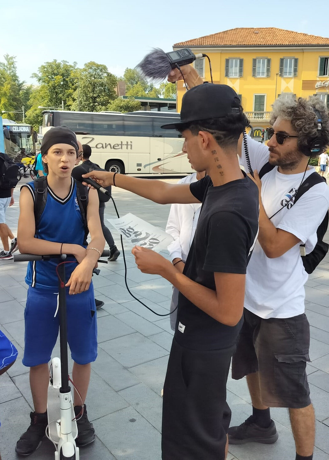 Un ragazzo ne sta intervistano un altro, tendendo verso di lui un microfono. Un altro ragazzo che indossa grandi cuffie sta registrando l'audio dell'intervista.
